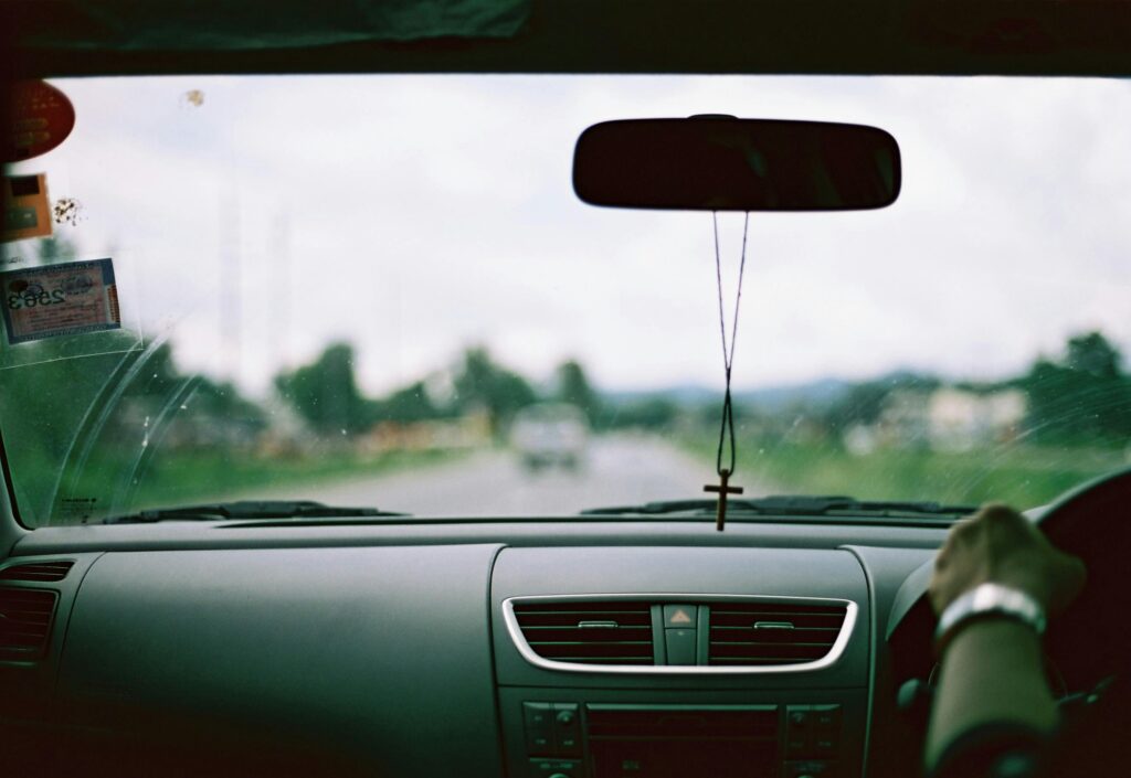 Interior view of a car dashboard with hanging cross on rear-view mirror, blurred road ahead.